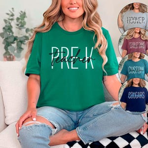 以下が含まれることがあります： 白と黒の文字で「PRE-K Teacher」と書かれた緑色のTシャツ。グレー、バーガンディ、ティール、ネイビーブルーのTシャツが追加で表示され、それぞれ異なるテキストが書かれています。その人はブルージーンズを履いています。