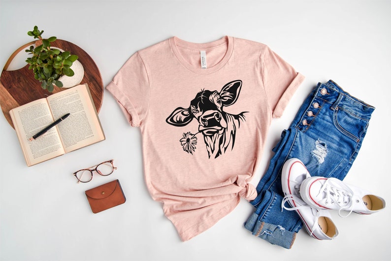 May include: A pink t-shirt with a black silhouette of a cow with a flower in its mouth. The shirt is laid out on a white surface with a pair of jeans, a book, a plant, glasses, and a wallet.