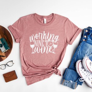 May include: A mauve t-shirt with the text "Working Wine to Wine" in white lettering. The text includes a clock and two wine glasses.