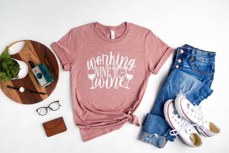 May include: A mauve t-shirt with the text "Working Wine to Wine" in white lettering. The text includes a clock and two wine glasses.