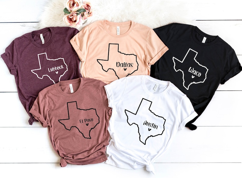 Op de afbeelding: Vijf T-shirts met een zwarte omtrek van de staat Texas en een stadsnaam die erin is gedrukt. De shirts zijn in verschillende kleuren, waaronder bordeauxrood, beige, zwart en wit. De genoemde steden zijn Lubbock, Dallas, Waco, El Paso en Austin.