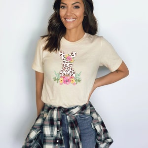 May include: A beige t-shirt with a floral leopard print Easter bunny graphic. The bunny is surrounded by pink, yellow, and green flowers.