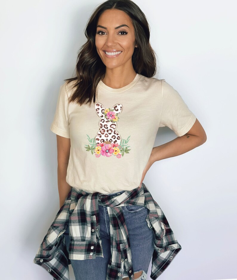 May include: A beige t-shirt with a floral leopard print Easter bunny graphic. The bunny is surrounded by pink, yellow, and green flowers.
