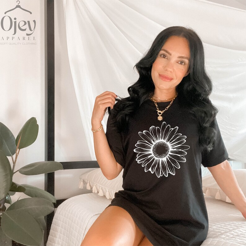 Puede incluir: Un vestido de camiseta negro con un gr&aacute;fico de margarita blanca. El vestido tiene mangas cortas y un ajuste holgado. La modelo lleva un collar de oro.