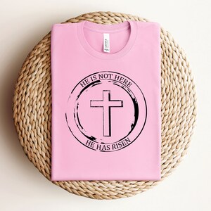 K&ouml;nnte beinhalten: Pinkes T-Shirt mit einem schwarzen Kreis-Motiv, auf dem "Er ist nicht hier, Er ist auferstanden" steht, mit einem Kreuz in der Mitte.
