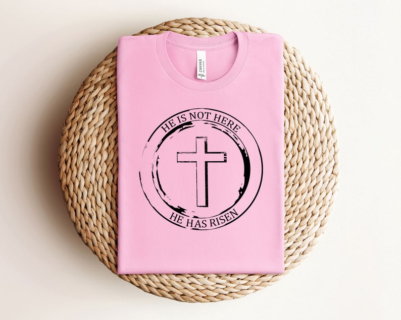 K&ouml;nnte beinhalten: Pinkes T-Shirt mit einem schwarzen Kreis-Motiv, auf dem "Er ist nicht hier, Er ist auferstanden" steht, mit einem Kreuz in der Mitte.