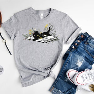 Puede incluir: Una camiseta gris jaspeada con un gato negro leyendo un libro rodeado de flores. El gato lleva una corona de flores. La camiseta est&aacute; sobre un fondo blanco con un par de vaqueros, una c&aacute;mara, gafas y un reloj.