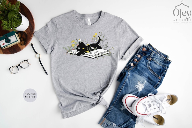 Puede incluir: Una camiseta gris jaspeada con un gato negro leyendo un libro rodeado de flores. El gato lleva una corona de flores. La camiseta est&aacute; sobre un fondo blanco con un par de vaqueros, una c&aacute;mara, gafas y un reloj.