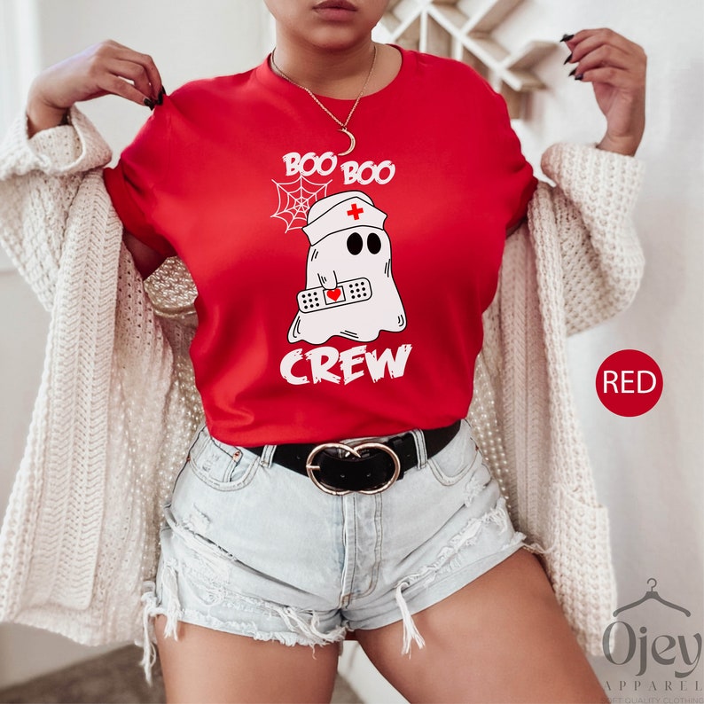 May include: Red t-shirt featuring a white ghost wearing a nurse's hat and holding a bandage. The text "Boo Boo Crew" is printed on the shirt.