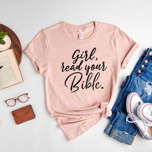 Pode incluir: Uma camiseta rosa com o texto "Girl, read your Bible." em escrita preta. A camiseta est&aacute; disposta sobre uma superf&iacute;cie branca com um livro, &oacute;culos, uma carteira, uma planta em vaso, uma cal&ccedil;a jeans e t&ecirc;nis brancos.