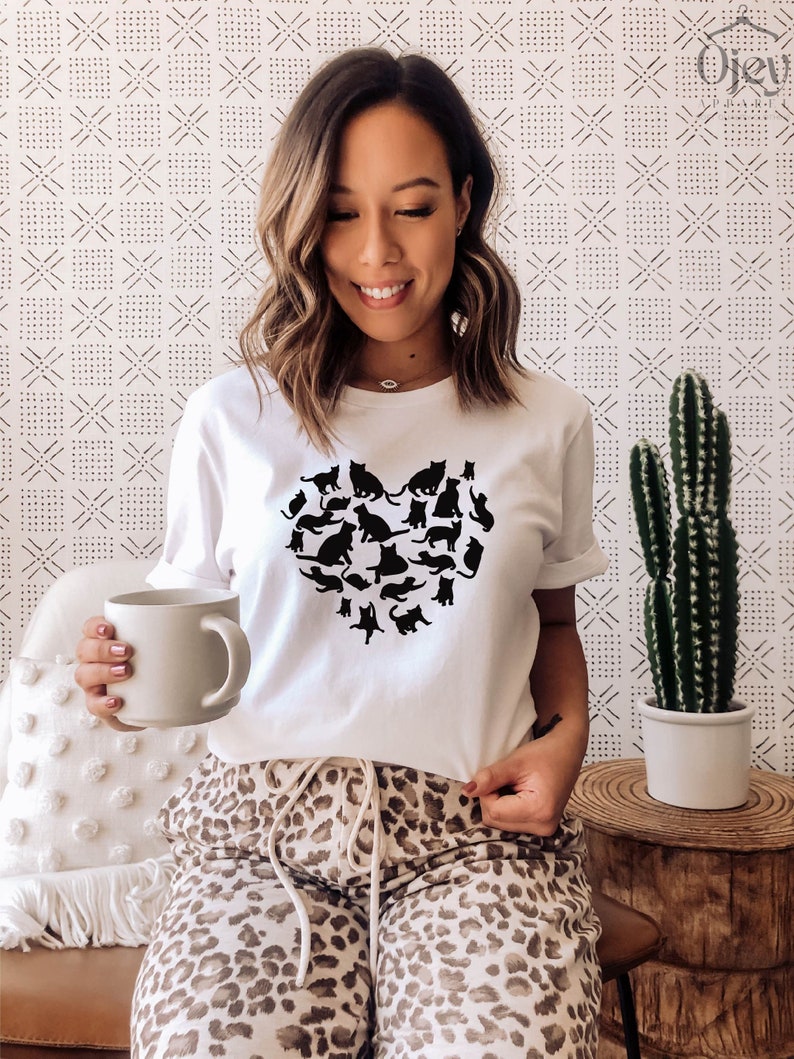 May include: A white t-shirt with a black silhouette of a heart made up of cats. The person wearing the shirt is sitting on a couch with a leopard print pattern.