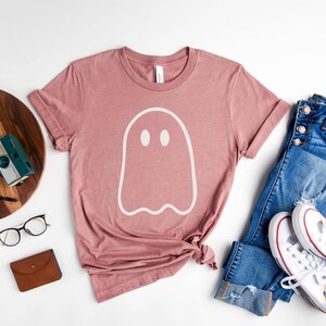May include: A heather mauve t-shirt with a white ghost graphic. The shirt is laid flat on a white surface with a pair of glasses, a wooden cutting board, a camera, a watch, a brown wallet, and a pair of blue jeans with white sneakers.