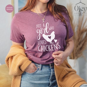 Puede incluir: Una camiseta de color marr&oacute;n morado con el texto "Just a girl who loves chickens" y una silueta blanca de una gallina con un coraz&oacute;n en su interior.
