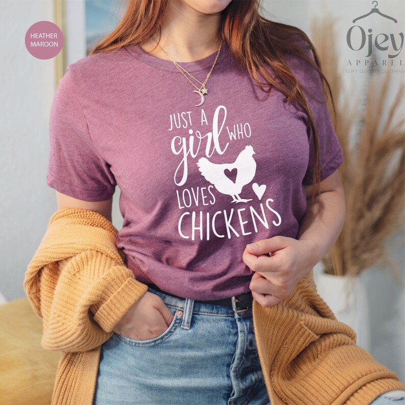 Puede incluir: Una camiseta de color marr&oacute;n morado con el texto "Just a girl who loves chickens" y una silueta blanca de una gallina con un coraz&oacute;n en su interior.