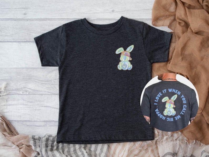Puede incluir: Una camiseta gris oscuro con un conejito arco&iacute;ris con un sombrero en la parte delantera. La parte posterior de la camiseta tiene un conejito arco&iacute;ris con un sombrero dentro de un c&iacute;rculo con el texto "Me encanta cuando me llamas big hoppa".