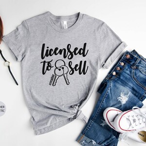 Pode incluir: Camiseta cinza com o texto "Licensed to Sell" e um par de chaves com a hashtag "#realtor" impressa nela. A camiseta &eacute; mostrada em um fundo branco com um par de jeans e t&ecirc;nis brancos.