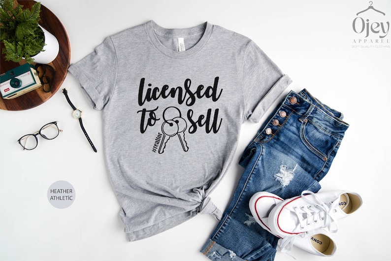 Pode incluir: Camiseta cinza com o texto "Licensed to Sell" e um par de chaves com a hashtag "#realtor" impressa nela. A camiseta &eacute; mostrada em um fundo branco com um par de jeans e t&ecirc;nis brancos.