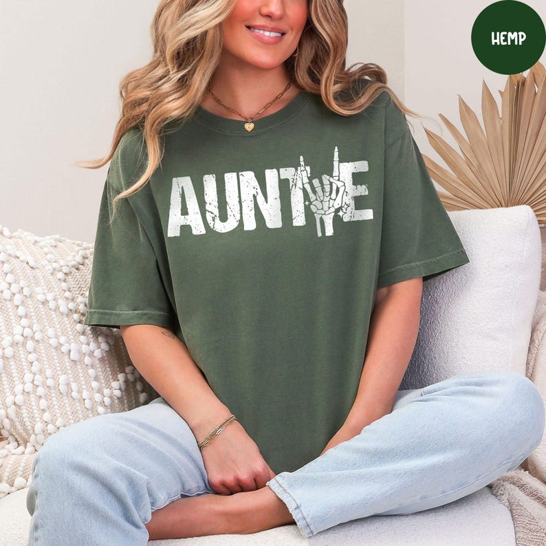 May include: Olive green t-shirt with the word "AUNTIE" in distressed white lettering. The "I" is replaced with a skeleton hand giving a peace sign. The shirt is made of hemp material.