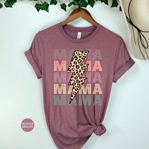 Op de afbeelding: Een heide-paarse t-shirt met een grafische afbeelding van een bliksem met een luipaardprint. De tekst "MAMA" wordt herhaald in een vervaagd, gelaagd effect in roze, oranje en grijs.