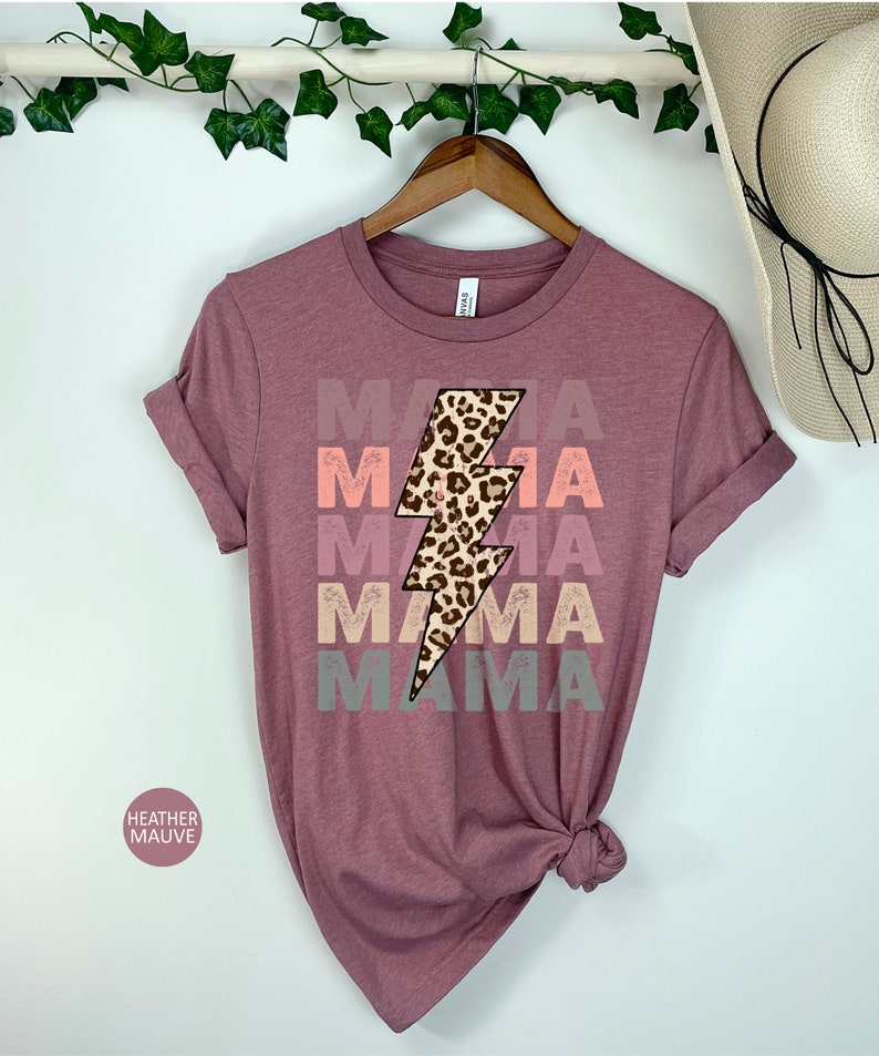 Op de afbeelding: Een heide-paarse t-shirt met een grafische afbeelding van een bliksem met een luipaardprint. De tekst "MAMA" wordt herhaald in een vervaagd, gelaagd effect in roze, oranje en grijs.