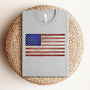 Pode incluir: Uma camiseta cinza com um design de bandeira americana desbotada estampado na frente.