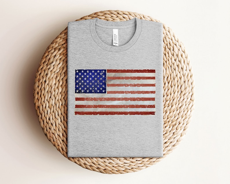 Pode incluir: Uma camiseta cinza com um design de bandeira americana desbotada estampado na frente.