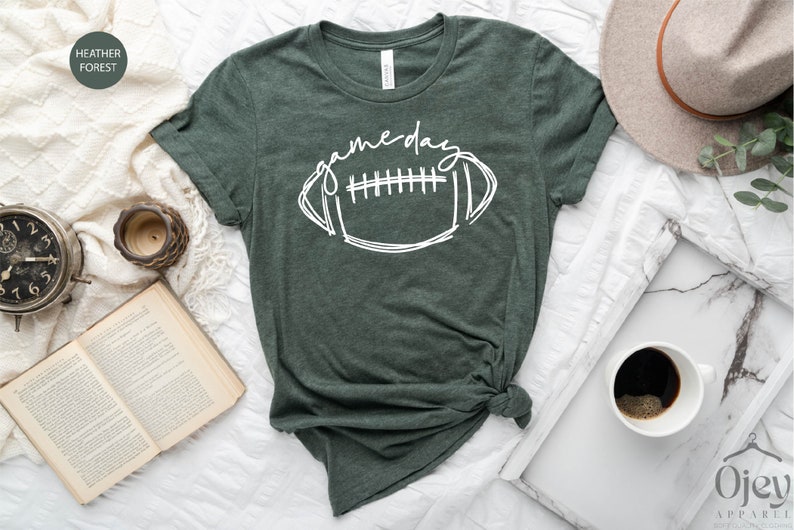 Peut inclure: Un t-shirt vert avec les mots "game day" et un contour blanc d'un ballon de football. Le t-shirt est nou&eacute; en bas.