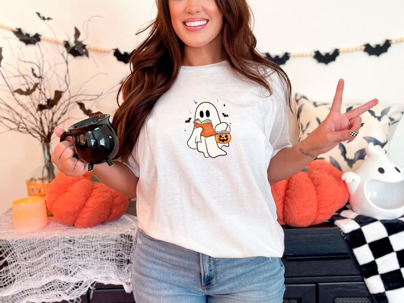 May include: White t-shirt with a graphic of a ghost holding a book and a pumpkin. The ghost is wearing a witch hat. The shirt is on a person wearing blue jeans.