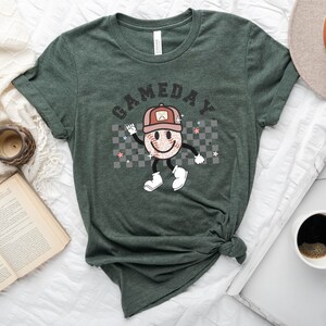 May include: A green t-shirt with a graphic of a smiling baseball with a hat and the text "GAMEDAY" printed on it.