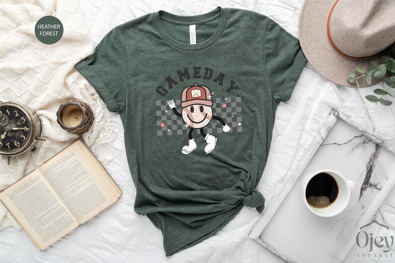 May include: A green t-shirt with a graphic of a smiling baseball with a hat and the text "GAMEDAY" printed on it.