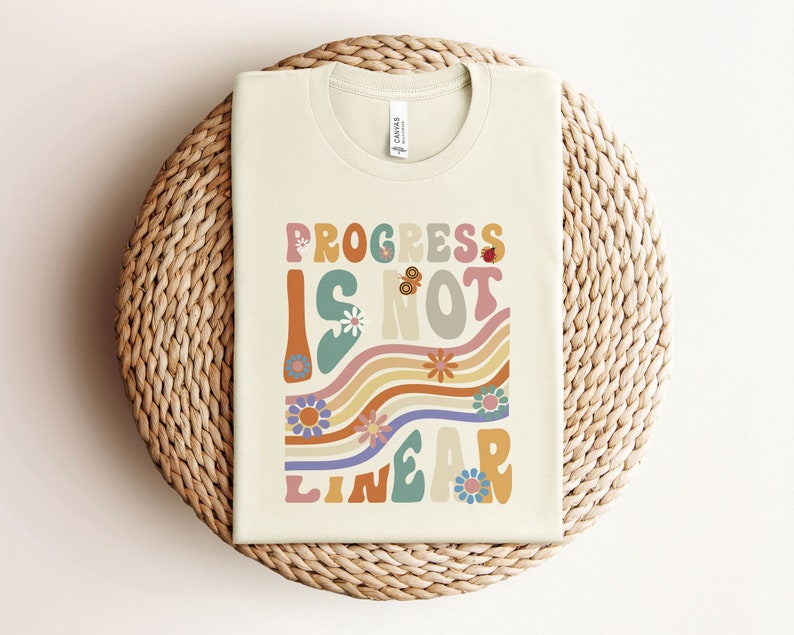 May include: A beige t-shirt with a retro-style graphic that says "Progress is not linear" in colorful, wavy letters. The graphic features flowers and butterflies.