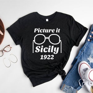 Pu&ograve; includere: Una maglietta nera con testo bianco che recita "Picture it Sicily 1922". Il testo &egrave; accompagnato da una grafica di un paio di occhiali da sole con una catena di perline.