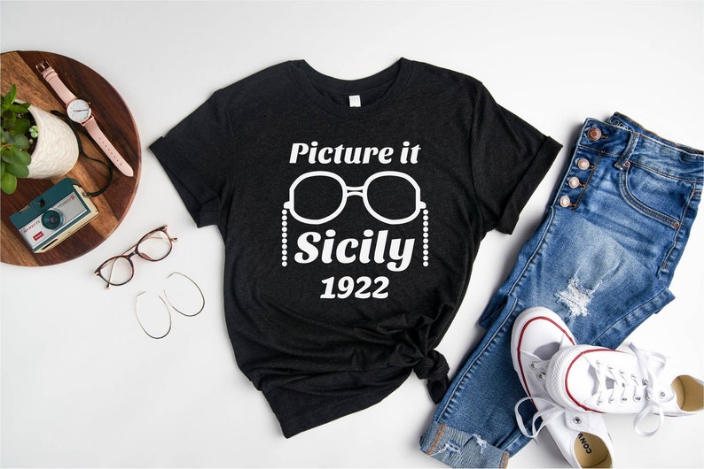 Pu&ograve; includere: Una maglietta nera con testo bianco che recita "Picture it Sicily 1922". Il testo &egrave; accompagnato da una grafica di un paio di occhiali da sole con una catena di perline.