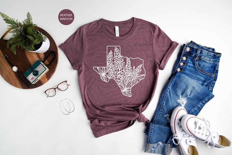 May include: Heather maroon t-shirt with a white graphic of the Texas state outline filled with floral designs. The shirt is styled with blue jeans and white sneakers. A wooden tray with a plant, camera, and accessories is also in the image.