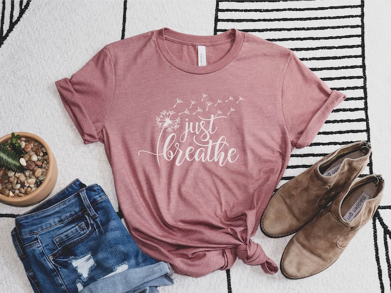K&ouml;nnte beinhalten: Ein mauvefarbenes T-Shirt mit dem Text "just breathe" in wei&szlig;en Buchstaben und einer L&ouml;wenzahndarstellung. Das Shirt ist gefaltet und auf einem wei&szlig;-schwarz gestreiften Teppich gelegt.