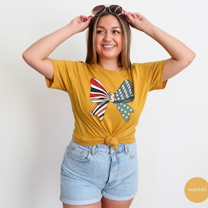 May include: A mustard yellow t-shirt with a red, white, and blue bow design. The bow is made of fabric with a vintage American flag pattern.