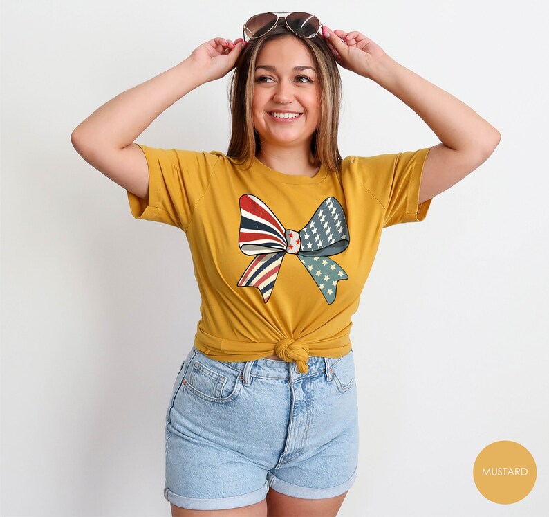 May include: A mustard yellow t-shirt with a red, white, and blue bow design. The bow is made of fabric with a vintage American flag pattern.