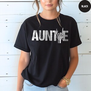May include: Black t-shirt with the word "AUNTIE" in distressed white lettering. The letter "E" is replaced with a skeleton hand making a rock and roll hand gesture. The shirt is a crew neck style.