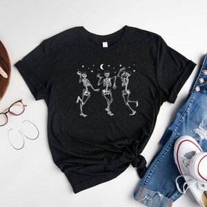 May include: A black t-shirt featuring a white graphic of three skeletons dancing beneath a crescent moon and stars. The t-shirt is laid out on a white surface with a pair of jeans, a watch, and a camera.