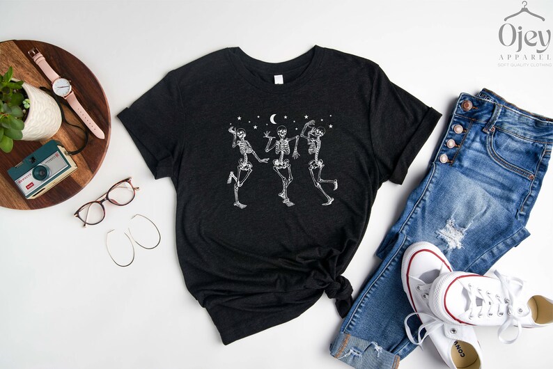 May include: A black t-shirt featuring a white graphic of three skeletons dancing beneath a crescent moon and stars. The t-shirt is laid out on a white surface with a pair of jeans, a watch, and a camera.