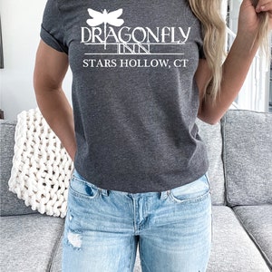 May include: A dark gray t-shirt with a white graphic of a dragonfly and the text "Dragonfly Inn Stars Hollow, CT".