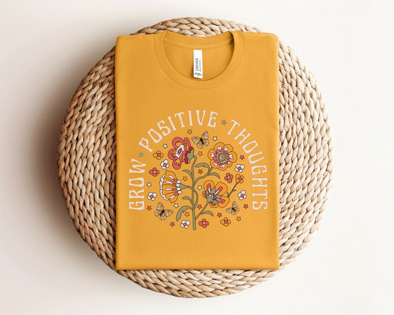 May include: A mustard yellow t-shirt with a floral design and the text "Grow Positive Thoughts" in a circle around the flowers.