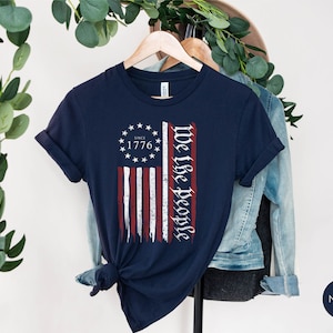 May include: Navy blue t-shirt with a distressed American flag design and the text "We the People" and "Since 1776".