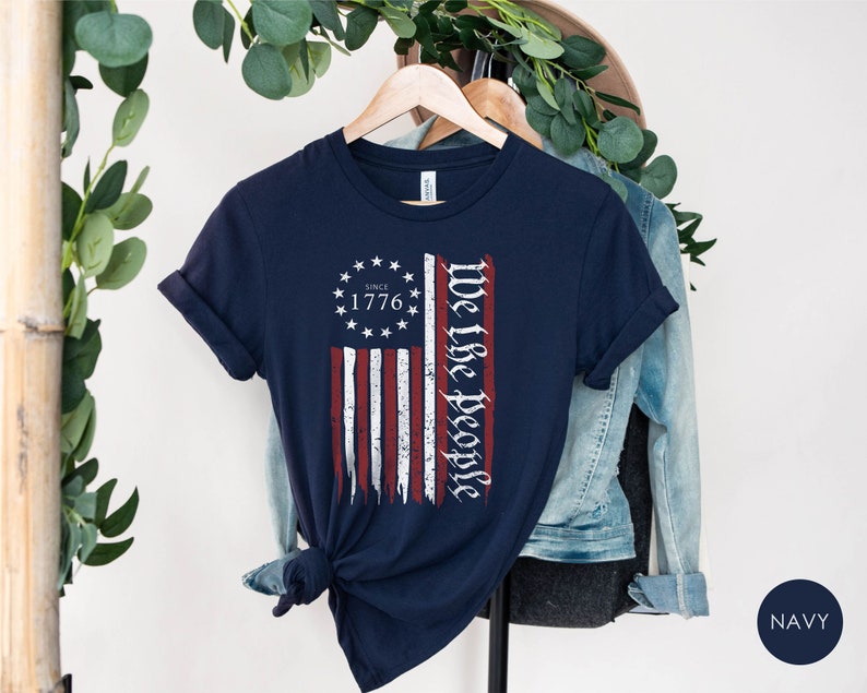 May include: Navy blue t-shirt with a distressed American flag design and the text "We the People" and "Since 1776".