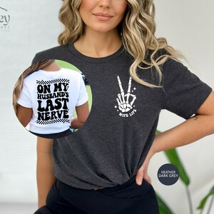 May include: A heather dark grey t-shirt with a skeleton hand making a peace sign and the text "Wife Life" on the front. The back of the shirt has the text "On My Husband's Last Nerve" in a retro font.