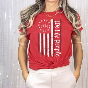 May include: Red t-shirt with a distressed American flag design and the text "We the People" printed on the front. The flag design includes the words "Since 1776" in a circle of stars.