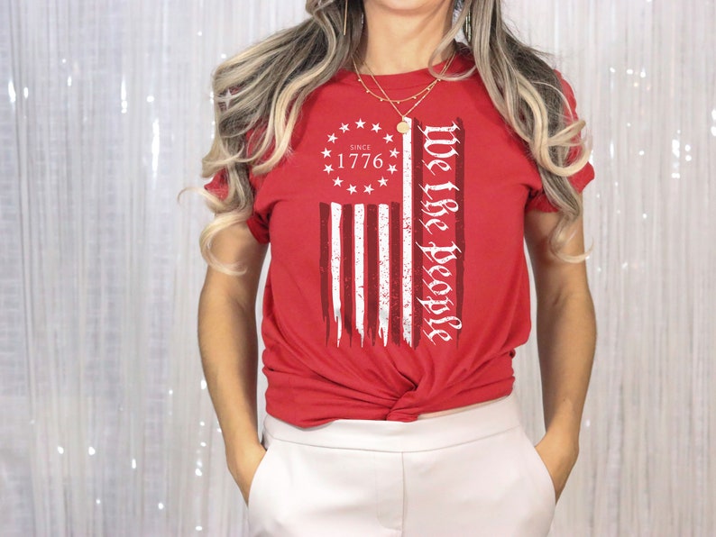 May include: Red t-shirt with a distressed American flag design and the text "We the People" printed on the front. The flag design includes the words "Since 1776" in a circle of stars.