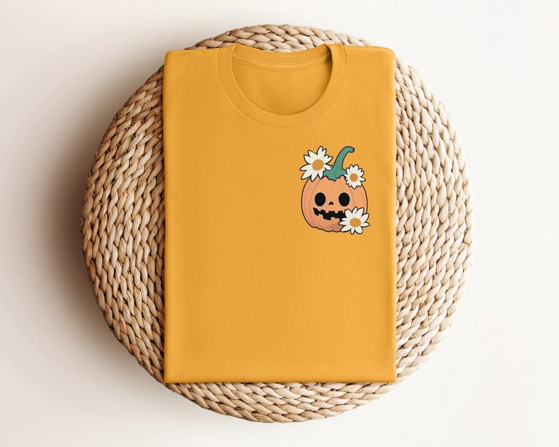 May include: A yellow t-shirt with a cartoon pumpkin wearing a flower crown. The pumpkin has a friendly face with a smile and eyes. The t-shirt is folded and displayed on a woven wicker surface.