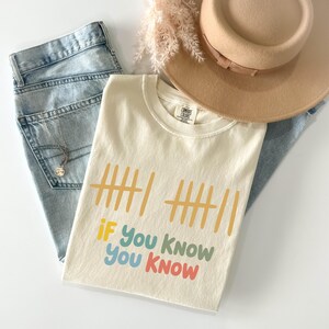 May include: Cream-colored t-shirt with the text "IF You KNOW You KNOW" in colorful letters. The shirt is folded and placed on top of light-wash denim jeans, a beige hat, and a necklace.