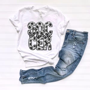 May include: White t-shirt with a black and white soccer ball design that says "Soccer Mama" on it. The shirt is paired with blue jeans.